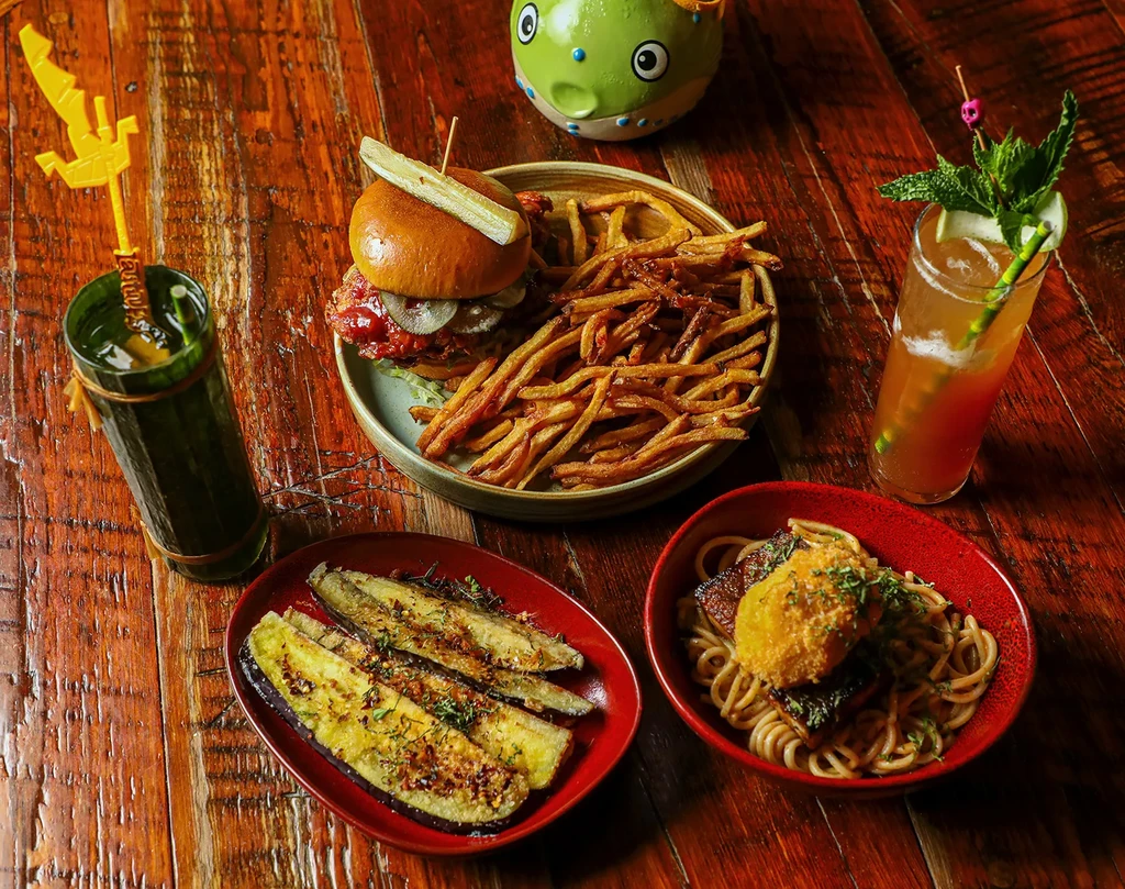 Overhead view of a wooden table with burger, noodles, eggplant, and drinks.