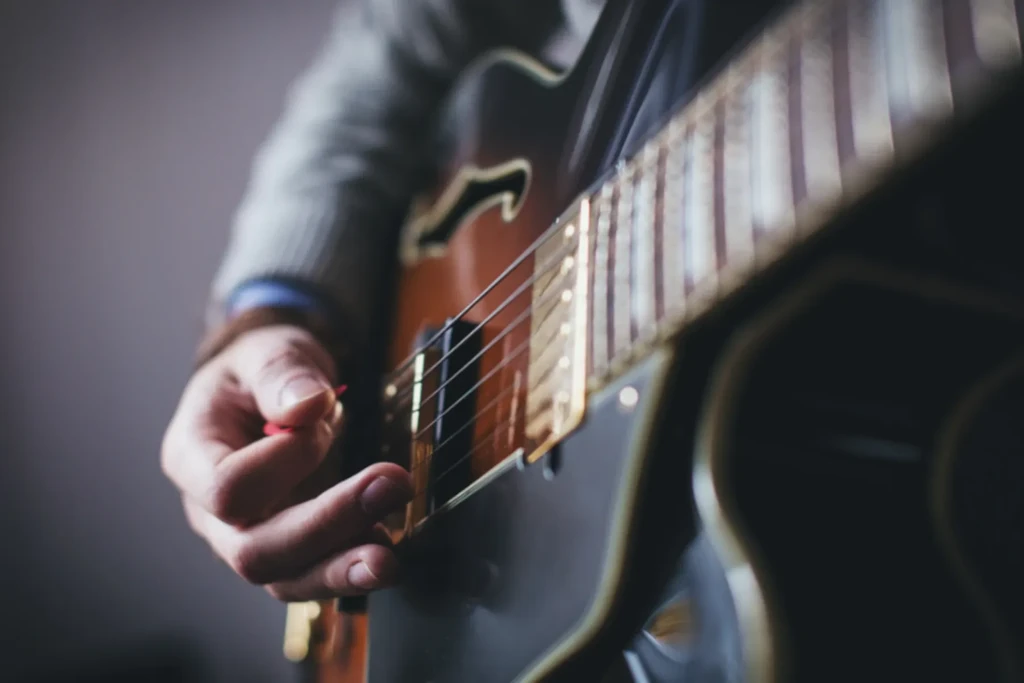 A hand with a red pick strums a brown guitar, showing golden hardware.
