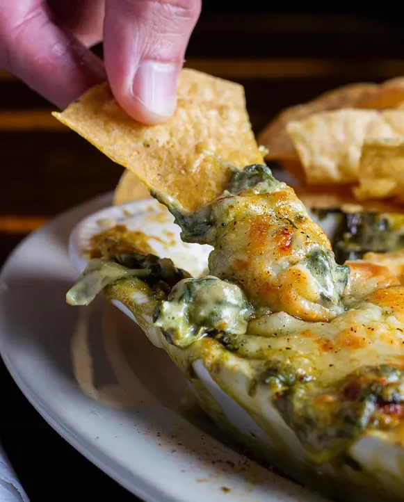 Hand dips a golden chip into warm, bubbly, cheesy green dip.