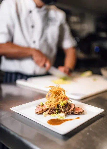 Plated seared tuna with garnishes in focus, chef works in blurred kitchen.