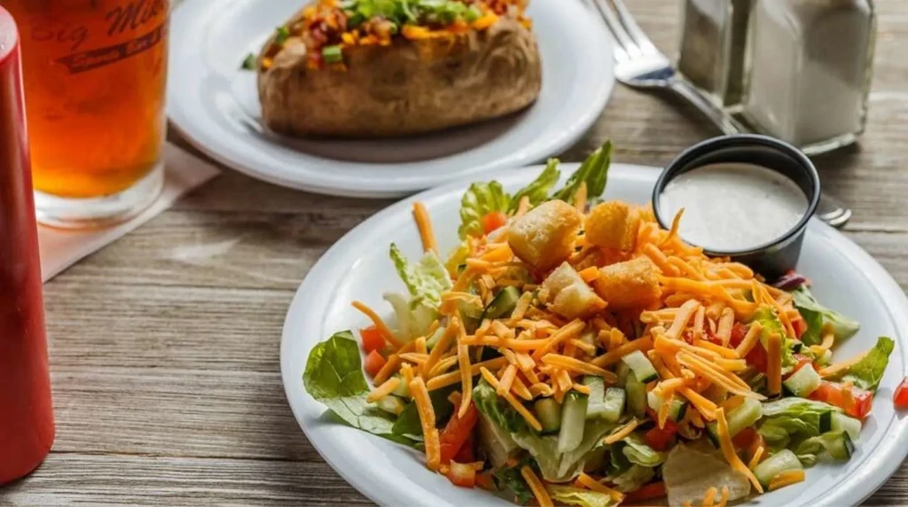 Green salad with cheddar, baked potato, and branded drink on wood table.