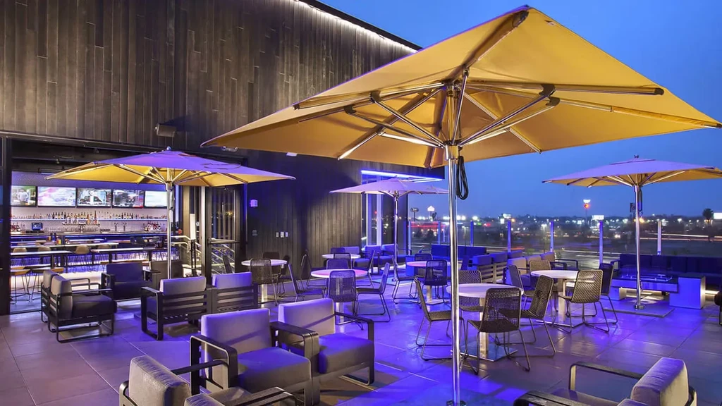 Nighttime rooftop patio with blue lights, yellow umbrellas, city skyline.
