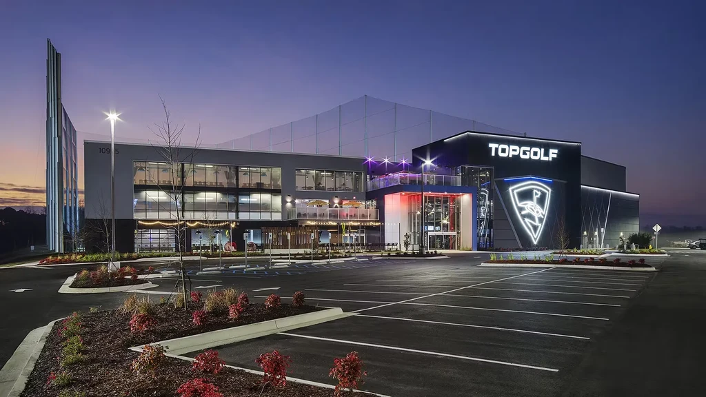 Modern Topgolf facility glowing at night with colorful lights and logo.