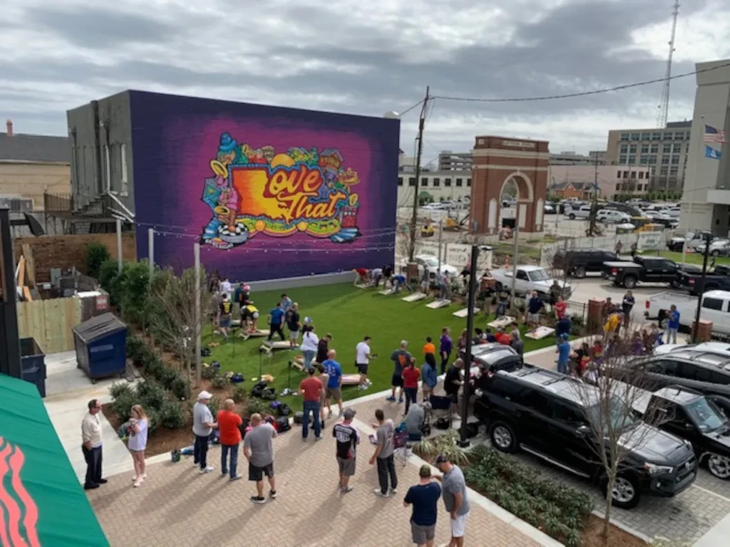 Outdoor urban gathering: people play cornhole on turf by a colorful mural.