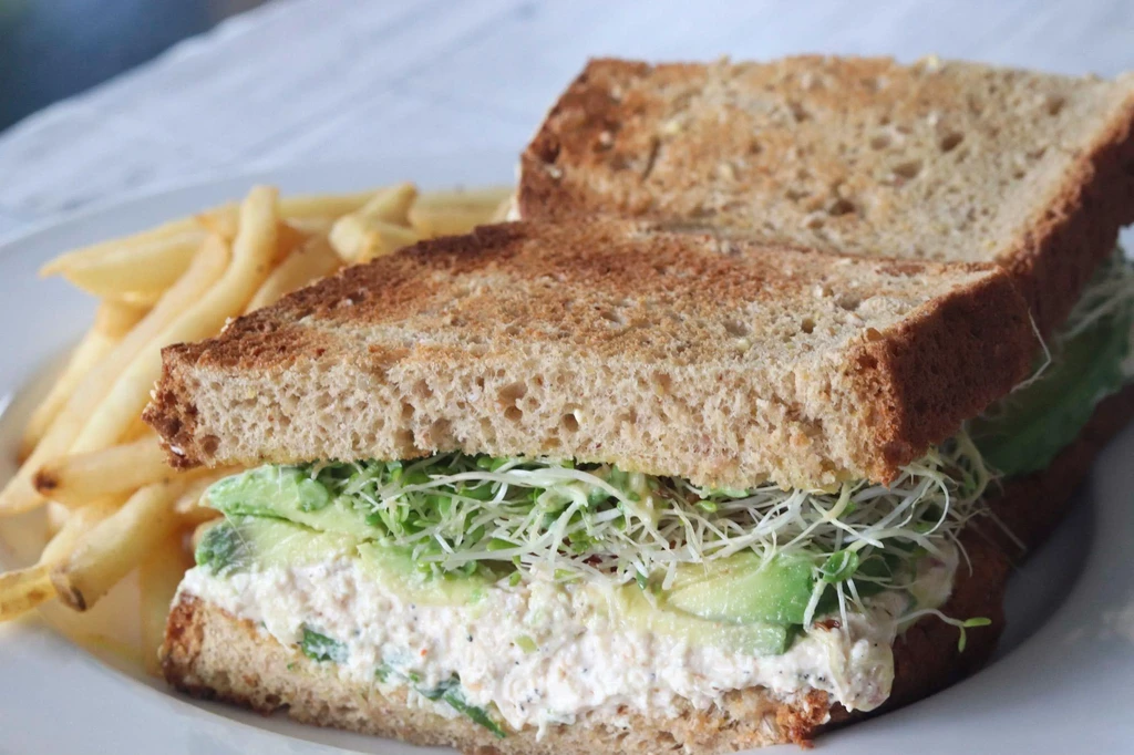 A toasted sandwich with creamy filling, avocado, sprouts, and fries.