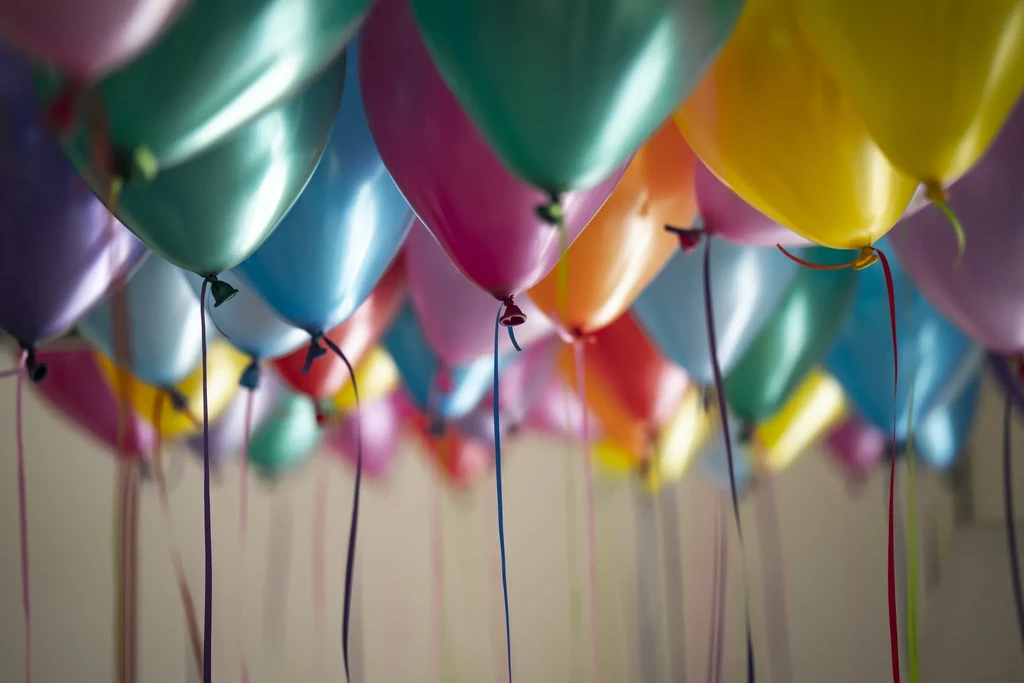 Numerous colorful, metallic balloons with ribbons float, filling the frame.