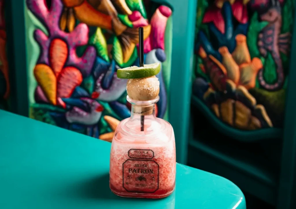 Reddish-pink frosty drink in miniature Patron bottle, lime, marine art.