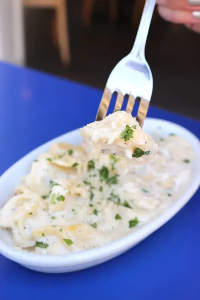 Fork lifts creamy dish with green herbs from white bowl on blue.