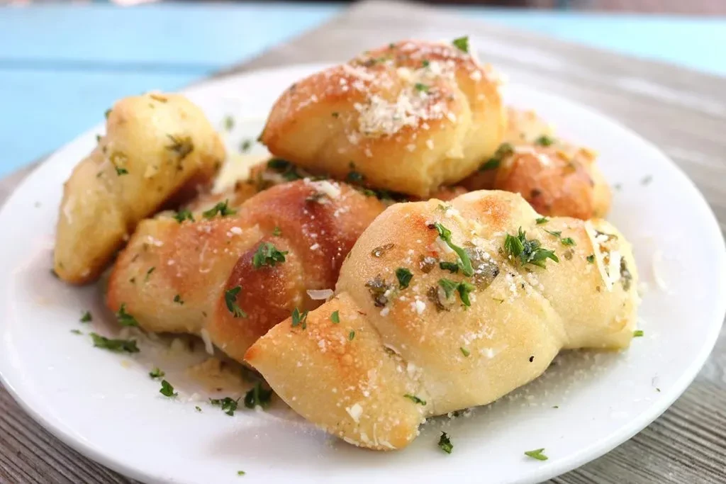 Golden-brown garlic knots with Parmesan cheese and parsley on a white plate.