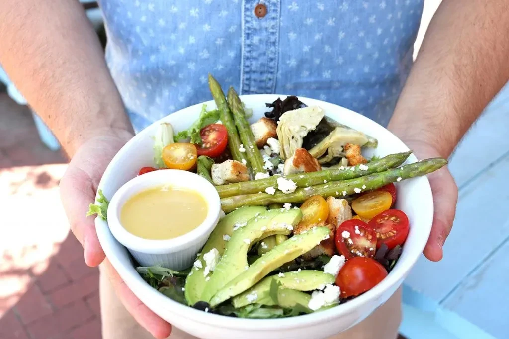 Person's hands holding a fresh salad with avocado, tomatoes, asparagus, and feta.