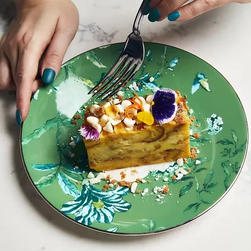 Hands hold a dessert with flowers on a jade-green botanical plate.