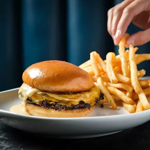 Close-up of double cheeseburger, golden fries on white dish, hand reaching.