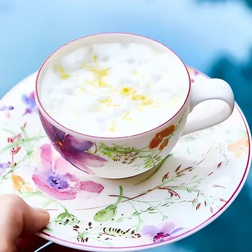 Hand holds floral teacup with foamy drink topped with yellow zest.