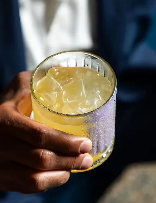 Dark-skinned hand holding a yellow drink with ice and purple glitter rim.