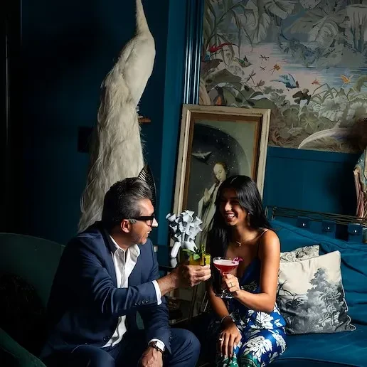 Two people clink cocktails on a teal couch, beside a peacock.