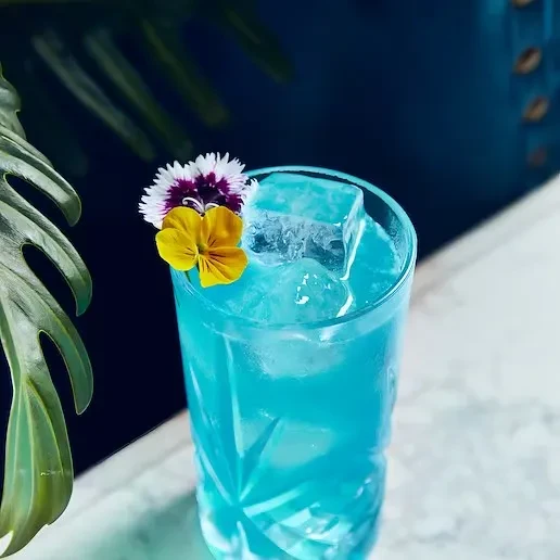 Vibrant blue cocktail with pansy, dianthus flowers, ice, and monstera leaf.