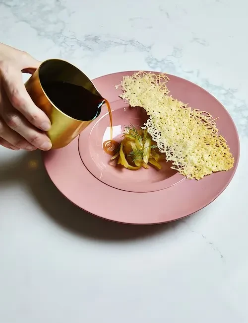 Hand pouring dark sauce over vegetables and crisp on a pink plate.