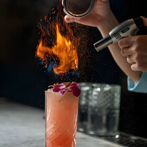Hands flame a pink cocktail with purple orchids at a dark bar.