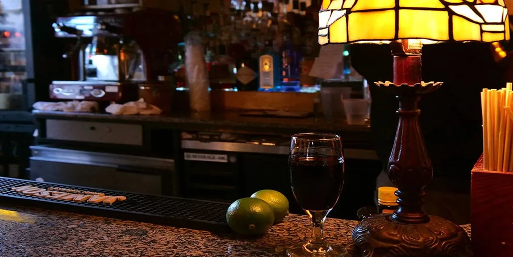 Warmly lit bar counter with cocktail, limes, ornate lamp, and blurred bottles.
