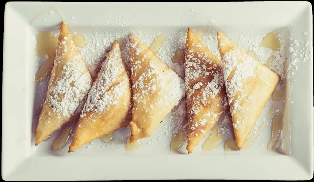 Five golden-brown triangular pastries dusted with sugar and drizzled with syrup.
