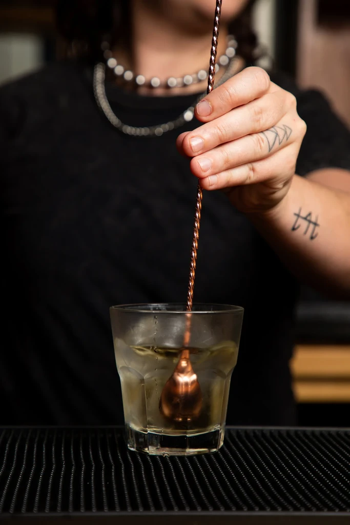 Tattooed hand stirs amber cocktail in rocks glass with copper spoon.