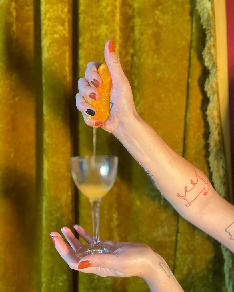Painted, tattooed hands squeeze orange juice into glass; velvet background.