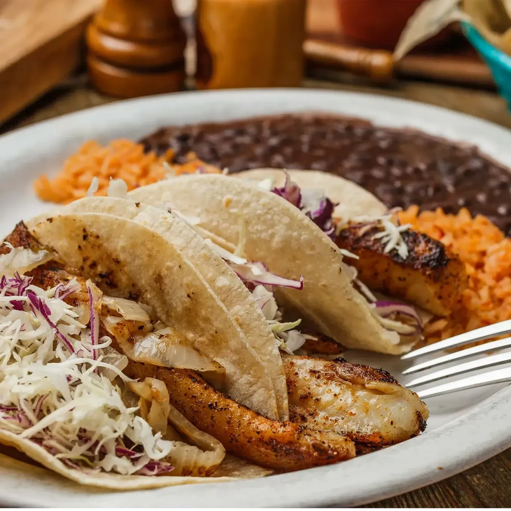 Plate of three grilled tacos, cabbage, rice, and black beans.