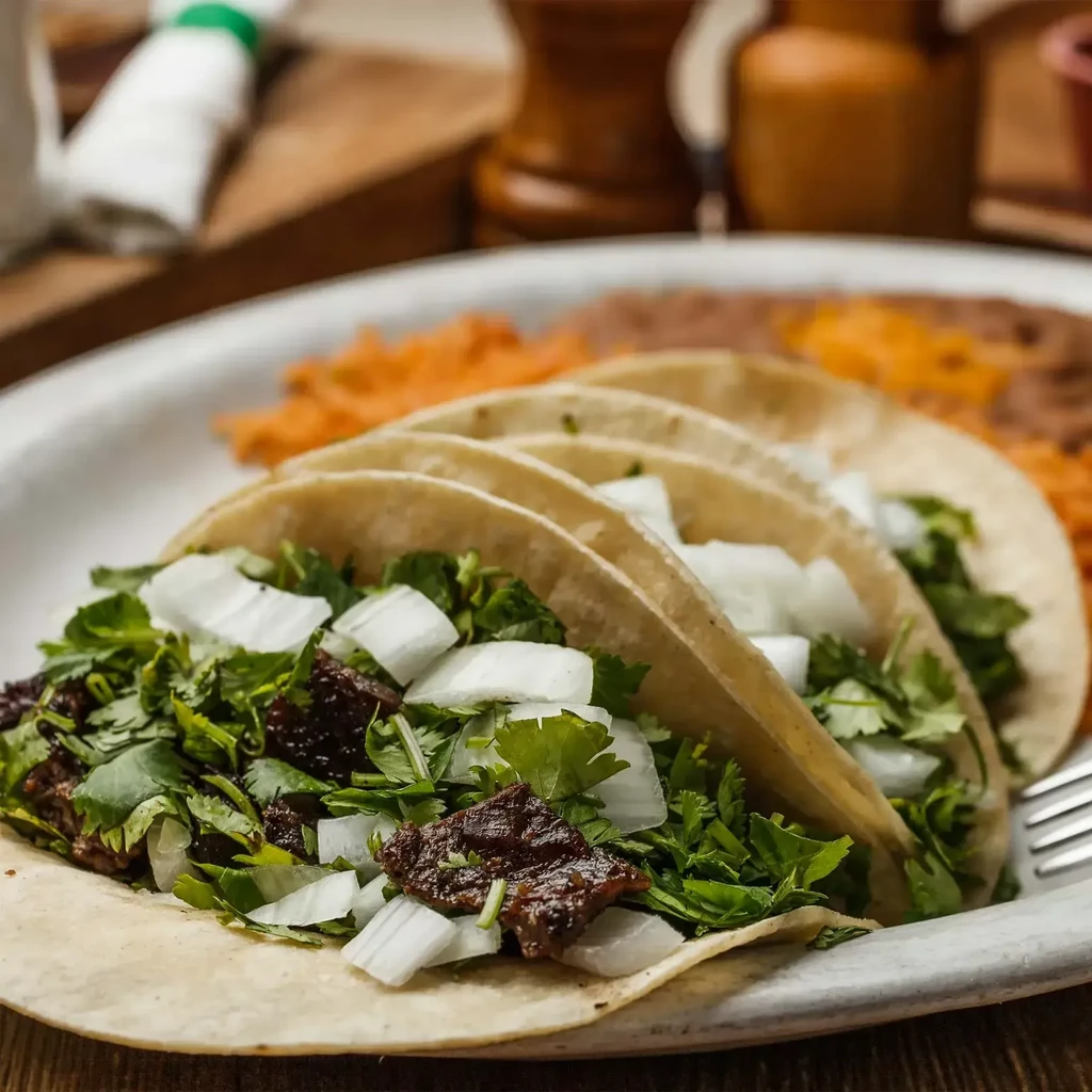 Plate of four beef tacos with cilantro, onions, rice, and beans.