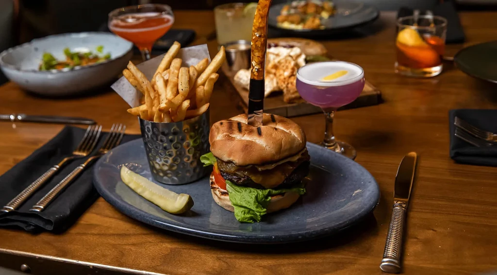 Gourmet burger with a knife, fries, pickle, and colorful cocktails.