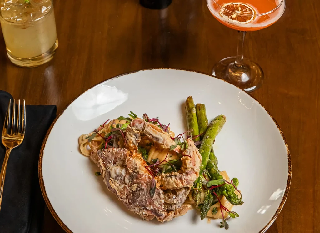 Fried soft-shell crab, pasta, asparagus, and two drinks on a dark table.