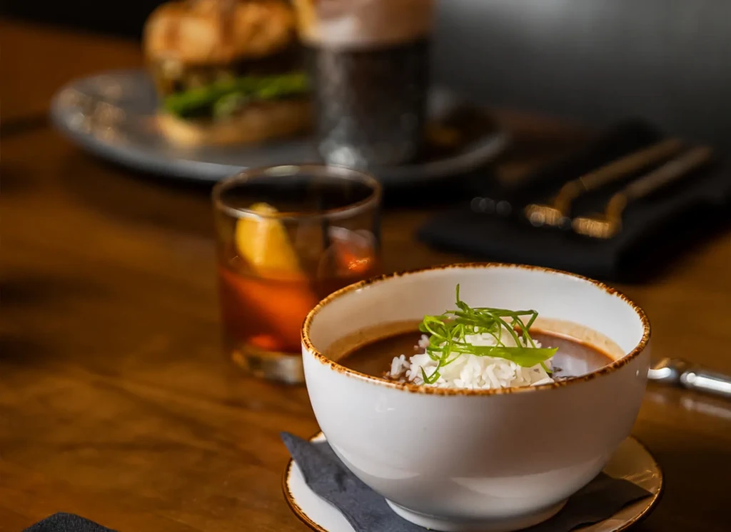 White bowl of dark soup with rice, drink, and blurred burger.
