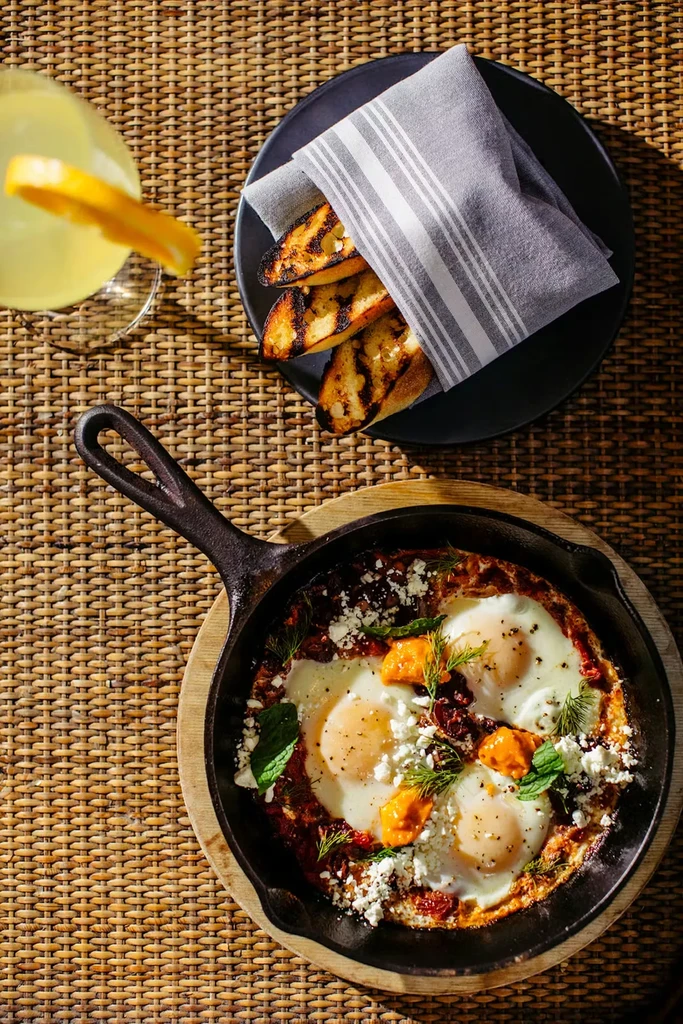 Shakshuka in cast iron pan, grilled bread, drink on wicker table.