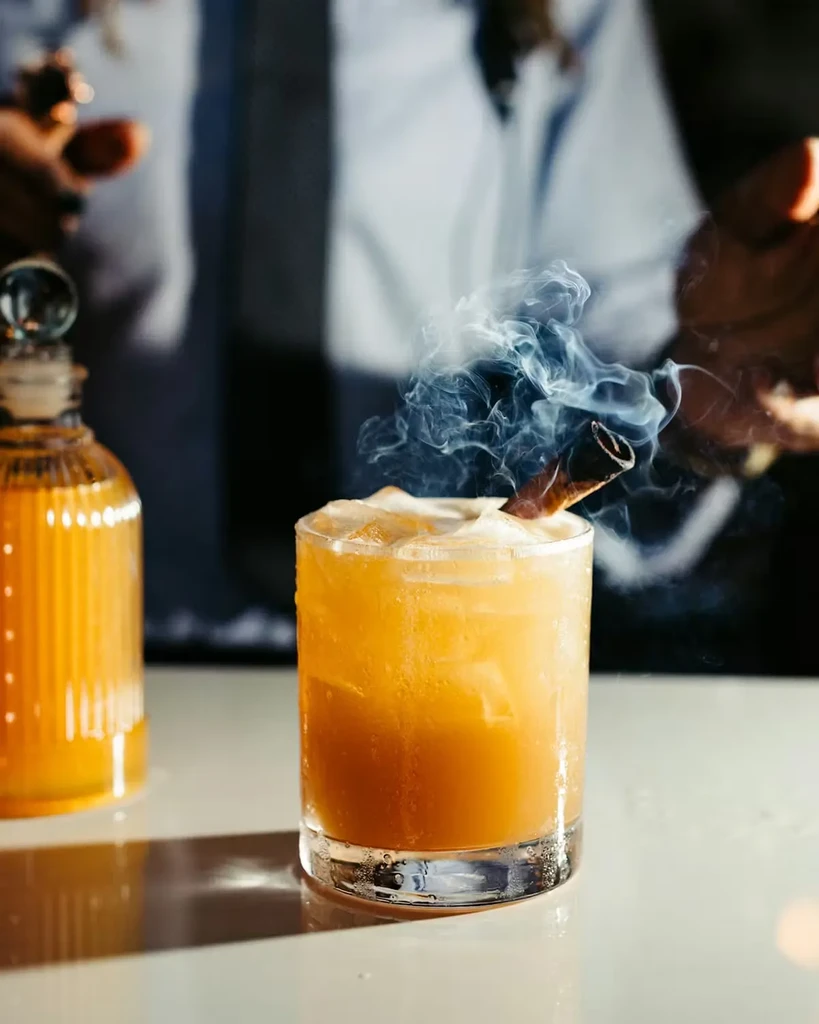 Amber cocktail with frothy top, ice, and smoking dark garnish.