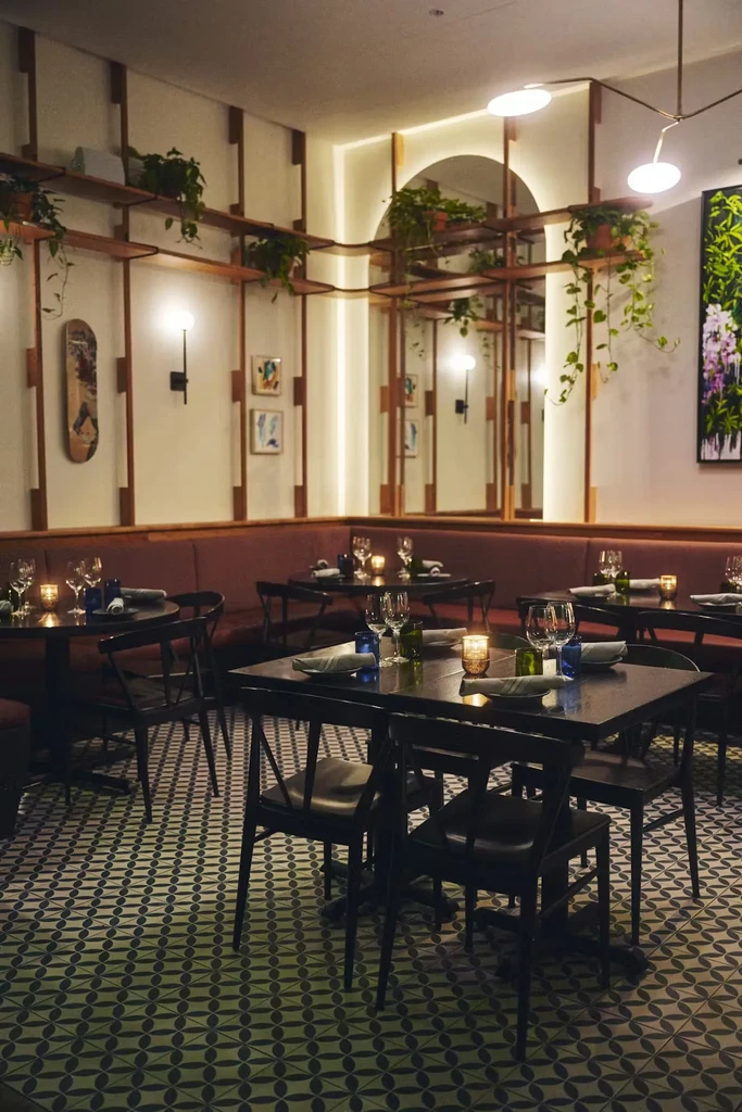 Warm, dimly lit restaurant interior with set tables, red banquette, and plants.