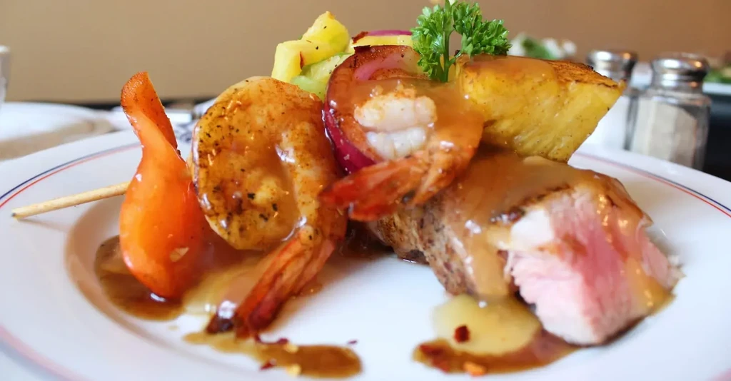 Gourmet surf-and-turf dish with shrimp, seared meat, sauce, and garnishes.