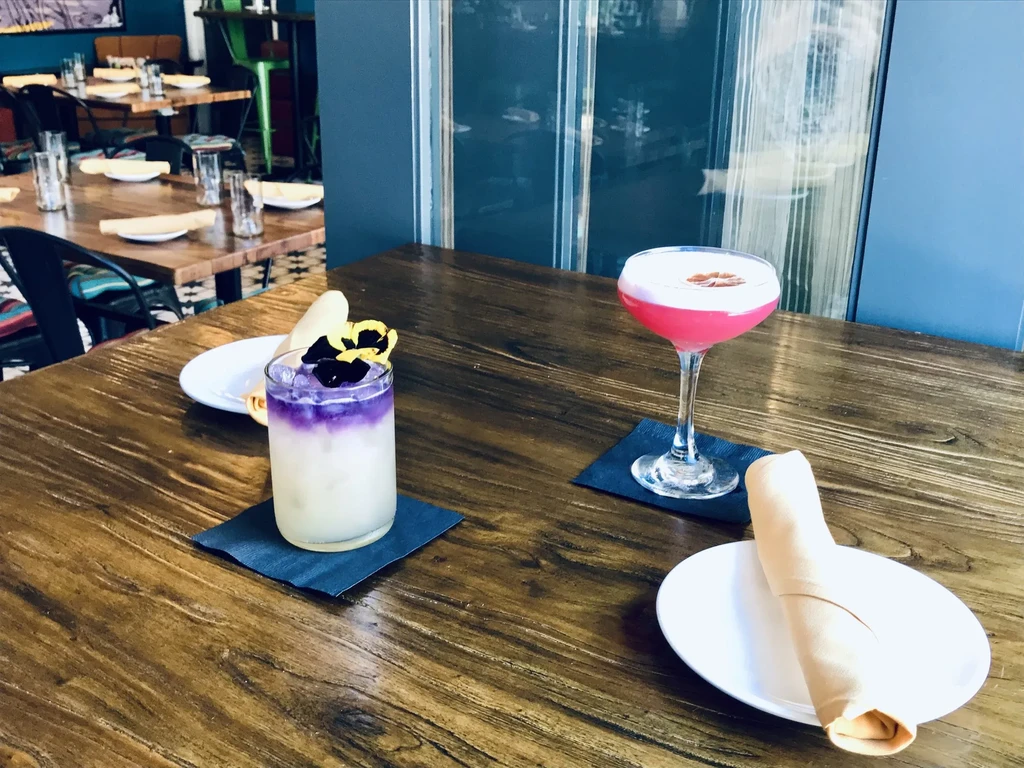 Purple flower-garnished and pink citrus-garnished cocktails on a wooden table.
