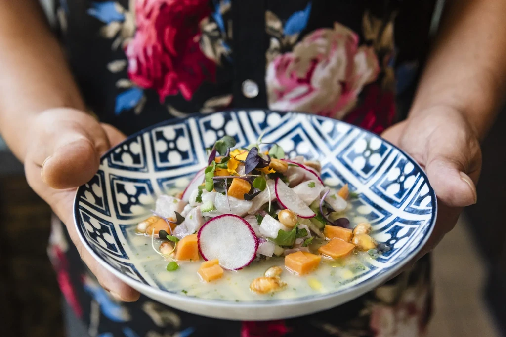 Hands holding vibrant ceviche with fish, radishes, sweet potato, and flowers.