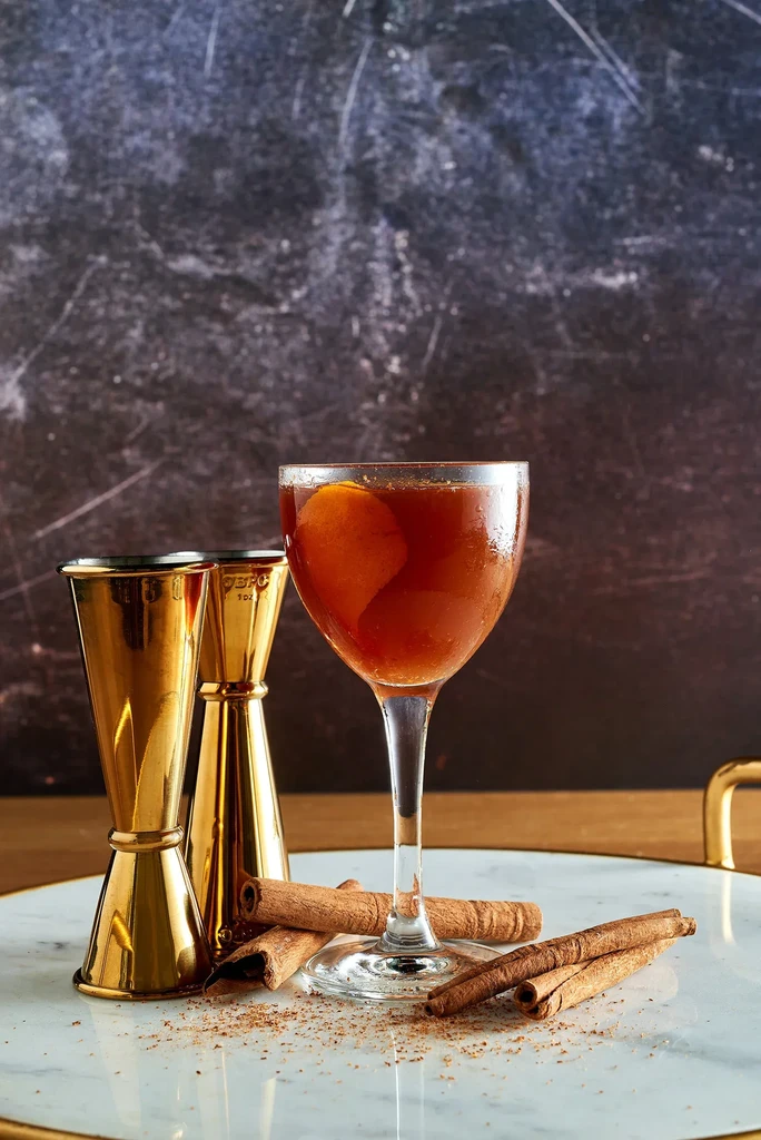 Dark amber cocktail with orange peel, brass jiggers, and cinnamon on marble.