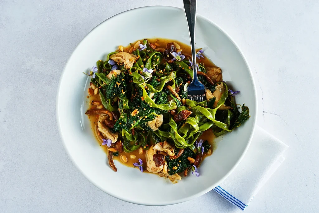 Plated green pasta with mushrooms, pine nuts, purple flowers, and sauce.