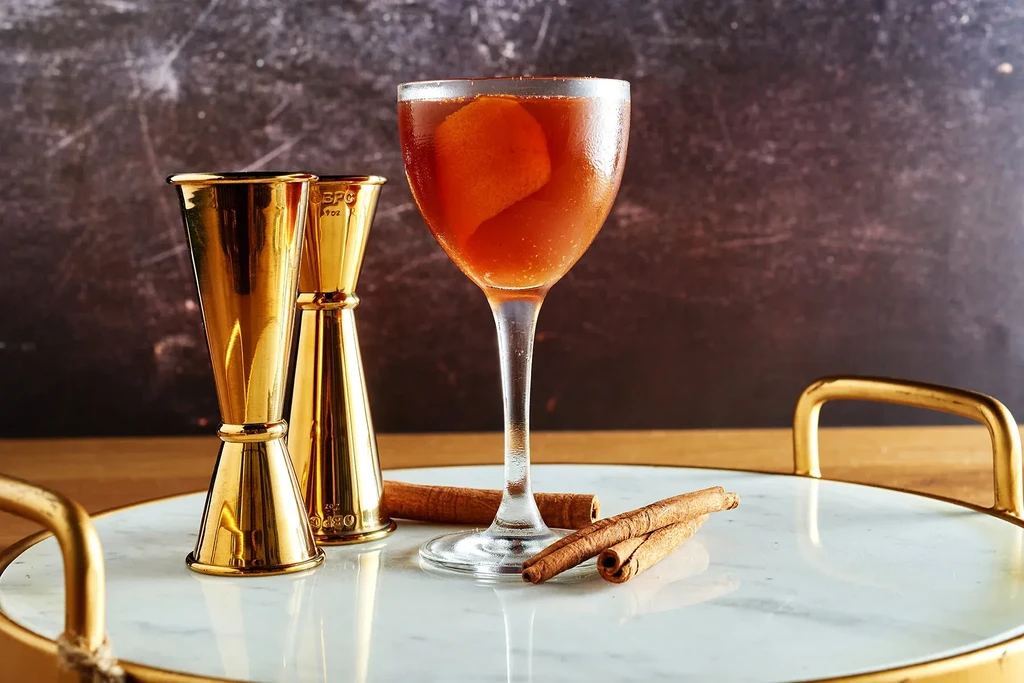 Chilled reddish-brown cocktail, orange peel, jiggers, and cinnamon on a marbled tray.