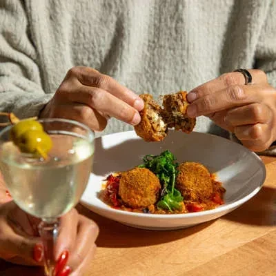 Hands breaking fried items with sauce, next to a garnished cocktail.