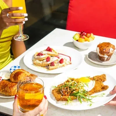 Vibrant tabletop with waffles, fruit, pastries, cutlet, and drinks; hands visible.
