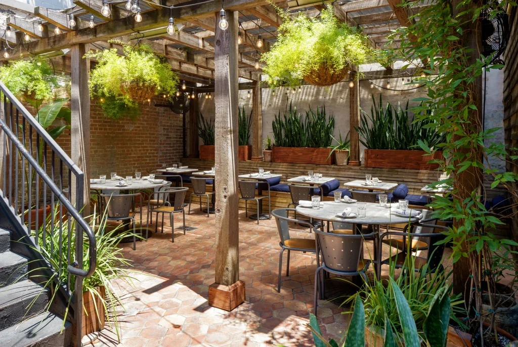 Empty outdoor dining patio with pergola, string lights, plants, and set tables.