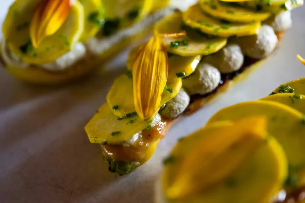 Gourmet dish with yellow slices, grey spheres, dark sauce, and a flower petal.