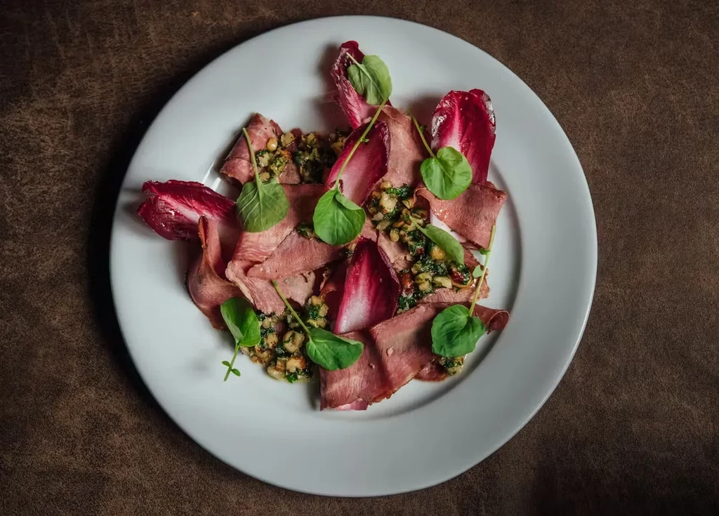 Finely plated sliced meat, crimson radicchio, green relish, and microgreens.