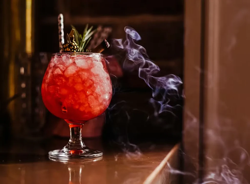 Vibrant red cocktail in stemmed glass with smoke and elegant garnishes.
