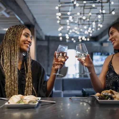 Two smiling women toast with wine glasses and food in a dim bar.