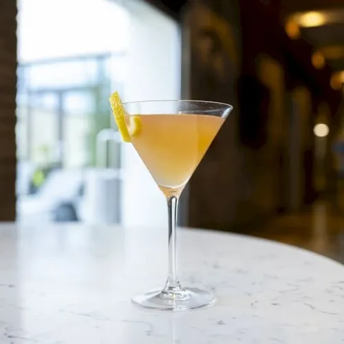 Pale orange martini cocktail with lemon peel on a white marble table.