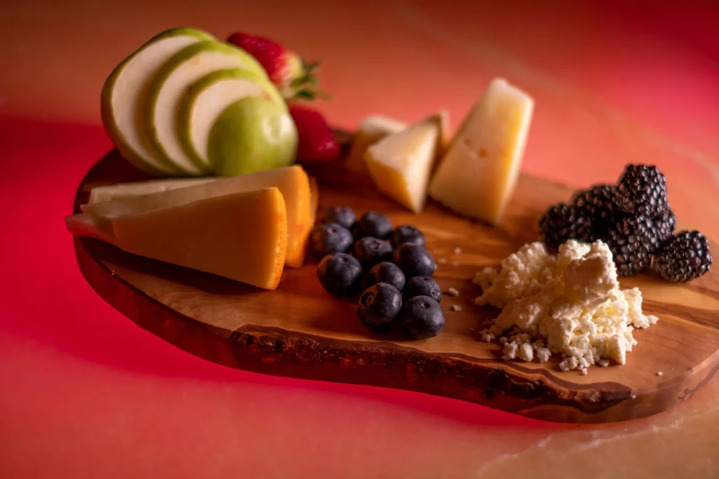 Rustic wooden board with various cheeses, fruits, and berries under red light.