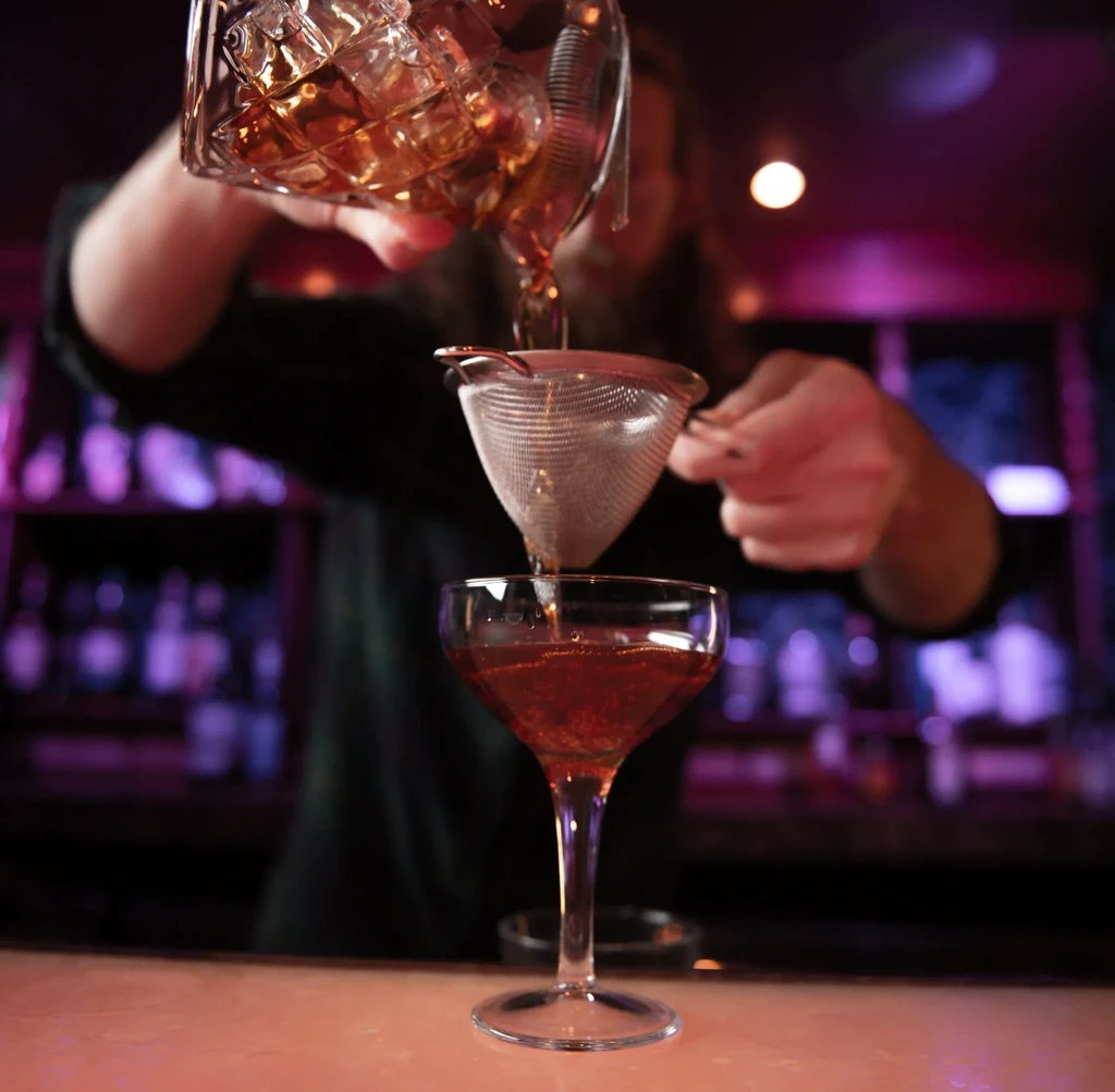 Bartender straining an amber cocktail from a mixing glass into a coupe.